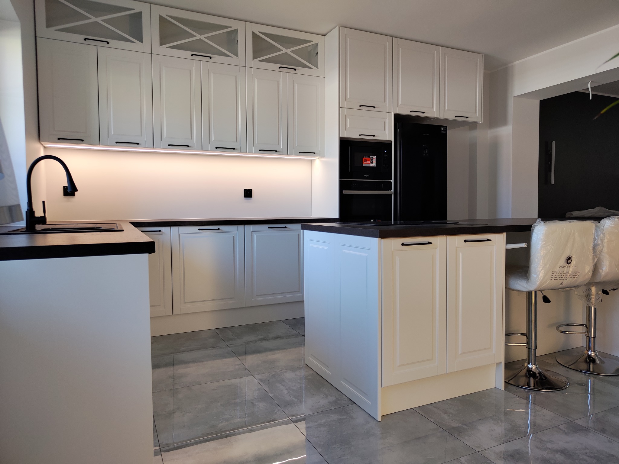 Nowoczesna, biała kuchnia z wyspą i czarnymi akcentami, oświetleniem LED pod szafkami i szarymi płytkami podłogowymi. Krzesła barowe z tworzywa sztucznego w folii ochronnej.
