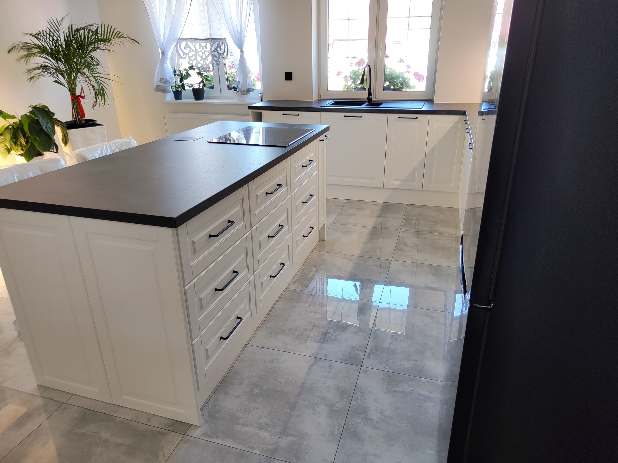 Biała kuchnia z wyspą i czarnym blatem, okno z kwiatami, szare płytki podłogowe. Minimalistyczny styl, nowoczesne wykończenie, roślina doniczkowa w tle.