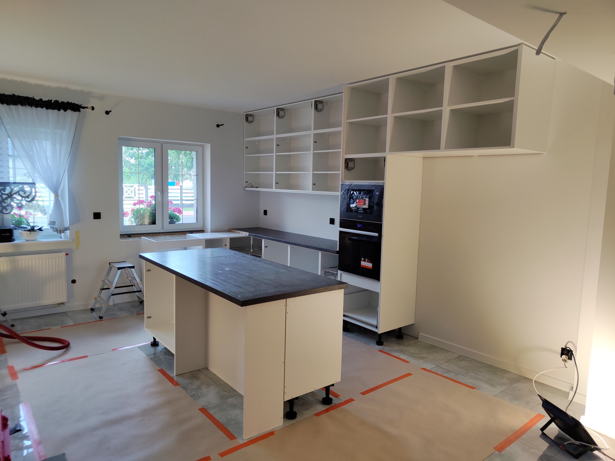 Montaż białej kuchni z wyspą i zabudową piekarnika. Widoczne górne szafki bez frontów, zabezpieczona podłoga. W tle okno z widokiem na zieleń.