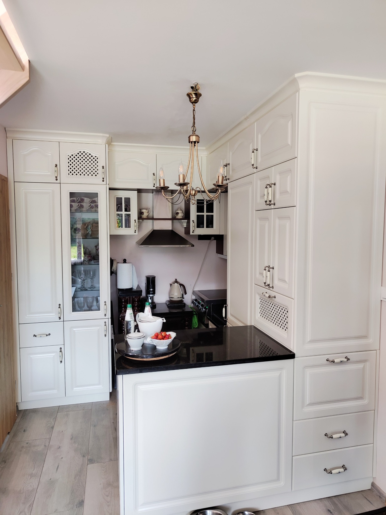 Biała, klasyczna kuchnia na wymiar z czarnym blatem. Widoczne przeszklone szafki i ozdobny żyrandol. Na blacie owoce i akcesoria kuchenne.