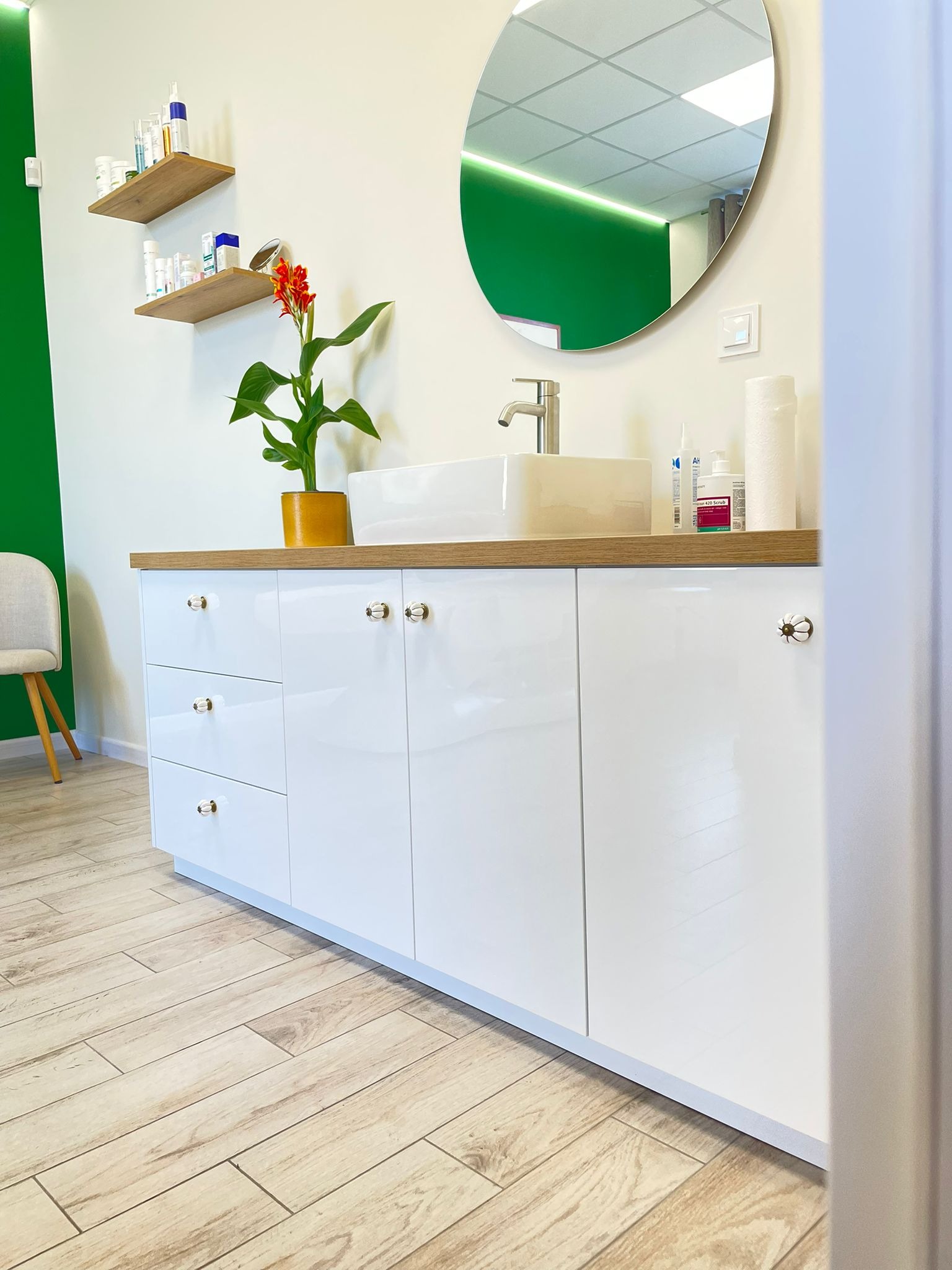 Biała szafka łazienkowa z blatem z drewna, okrągłe lustro, kwiat w doniczce i kosmetyki. Całość w nowoczesnym stylu.