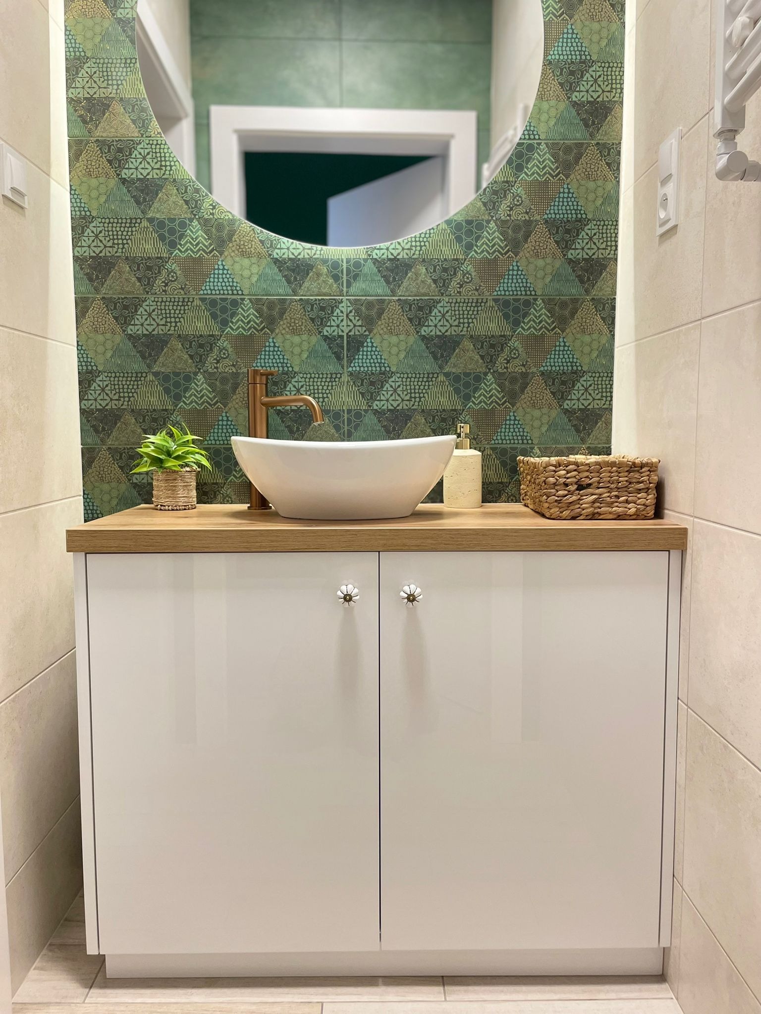 Biała szafka łazienkowa z umywalką nablatową i złotą armaturą. Nad nią lustro w geometryczne wzory. Dekoracje: roślina w doniczce i pleciony koszyk.