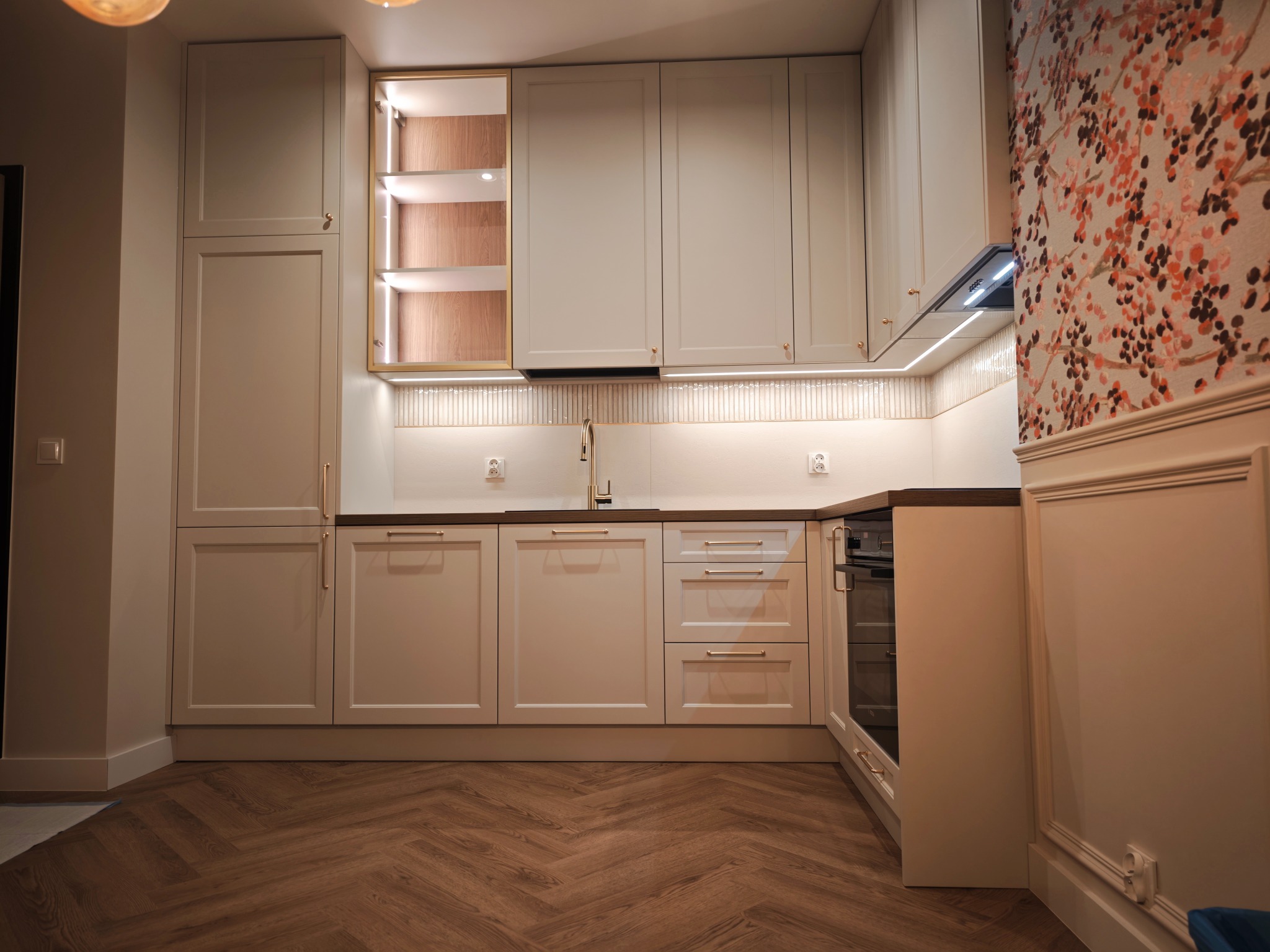 Jasna zabudowa kuchenna z oświetleniem LED i drewnianym blatem w Warnicach. Fronty frezowane, złote uchwyty, parkiet w jodełkę i ściana w kwiaty.