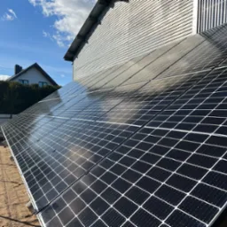 Panele fotowoltaiczne zamontowane na ziemi obok budynku z blachy falistej, w tle niebo z chmurami i dom jednorodzinny.