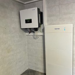 DKJM Install - Zamontowany na ścianie biały falownik Sofar z podłączonymi kablami oraz biały akumulator energii Sofar stojący obok w pomieszczeniu z wykafelkowanymi ścianami.