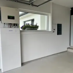 Biały magazyn energii SOFAR z inwerterem na ścianie w garażu, okno z kwiatami, szara podłoga i listwa przypodłogowa. Estetyczny montaż w jasnym wnętrzu.