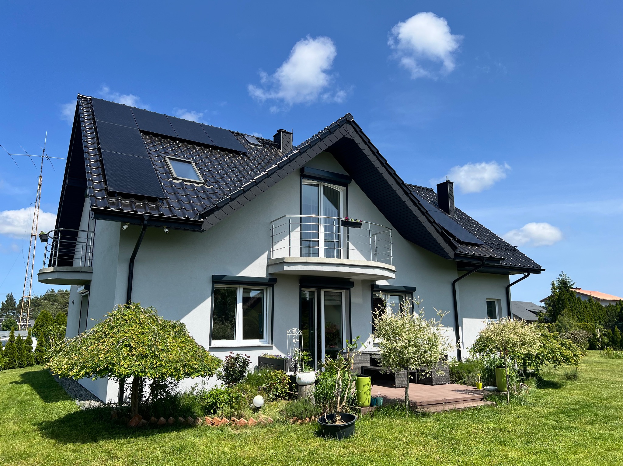 Dwupiętrowy dom z panelami fotowoltaicznymi na dachu, balkonem z metalową barierką i zadbanym ogrodem w słoneczny dzień. Widoczna antena na dachu.