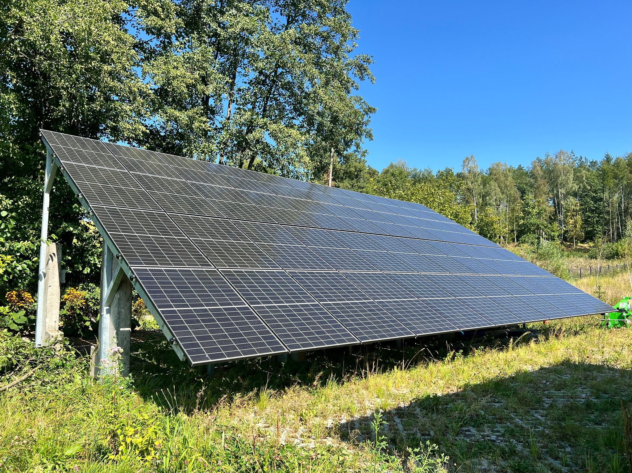 Panel fotowoltaiczny ustawiony na łące z widocznymi drzewami w tle, prezentujący instalację w naturalnym otoczeniu.