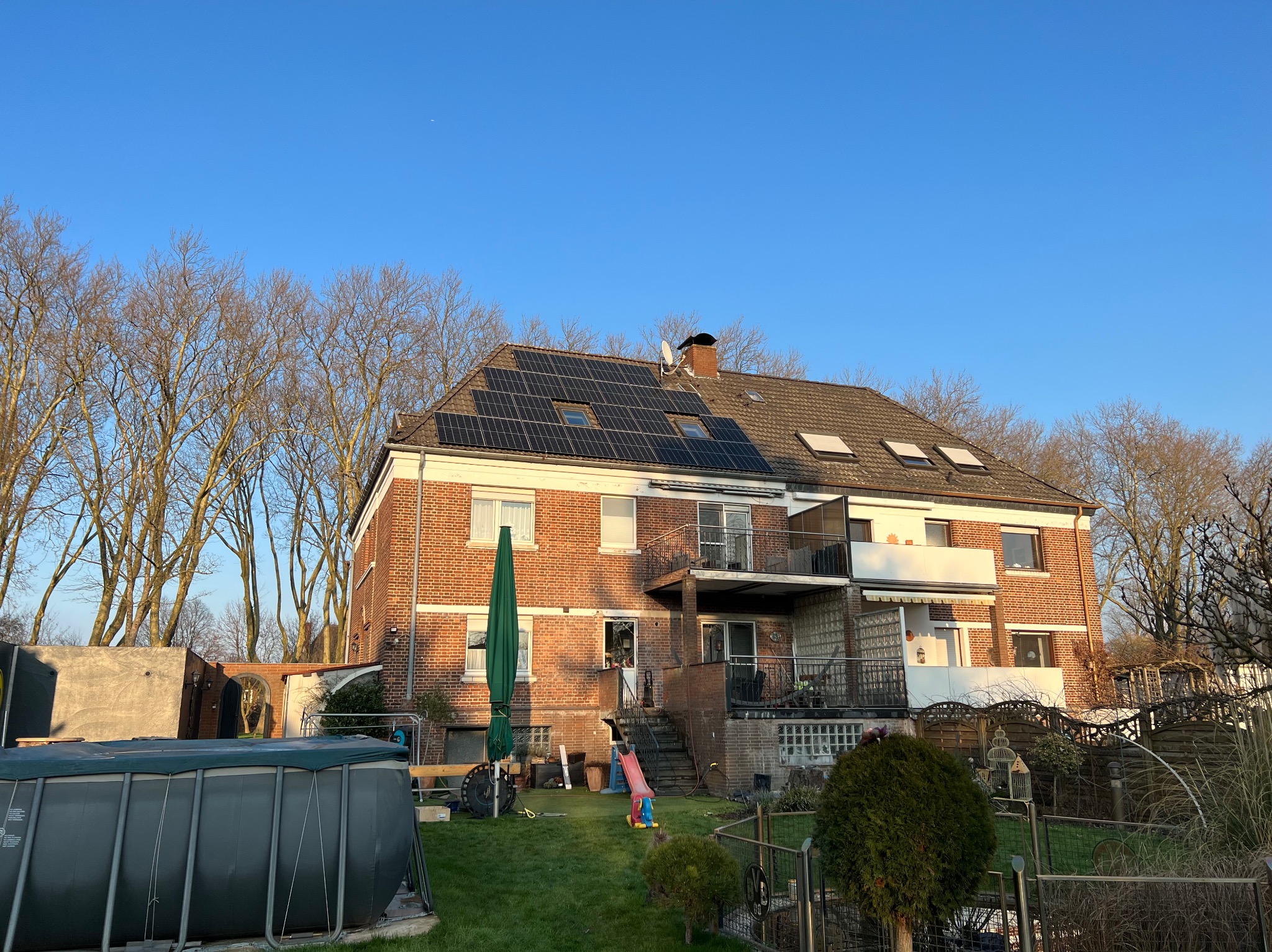 Ceglany dom z panelami fotowoltaicznymi na dachu, widok z ogrodu w słoneczny dzień. Na pierwszym planie basen i zieleń, w tle drzewa bez liści.