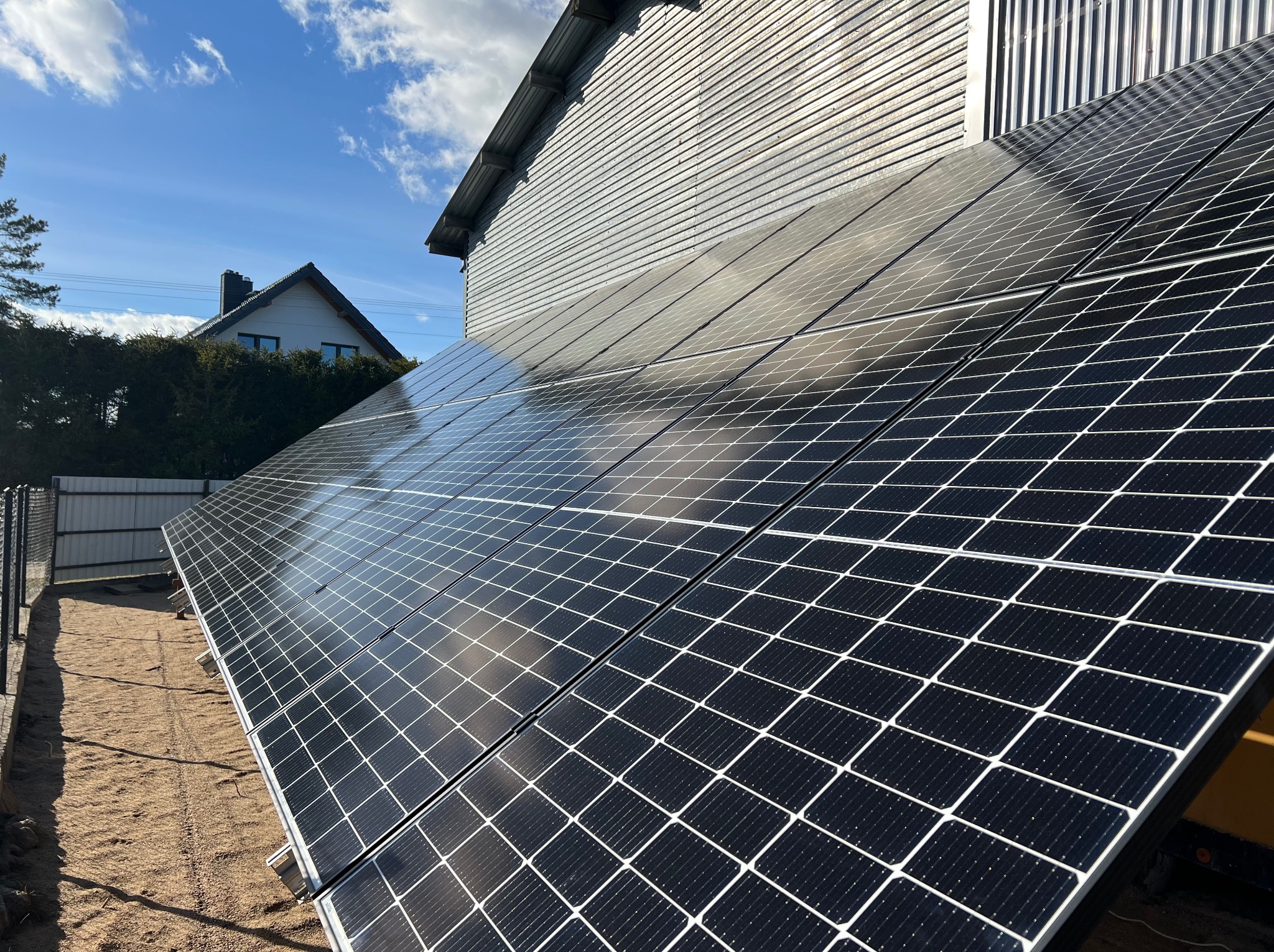 Panele fotowoltaiczne zamontowane na ziemi obok budynku z blachy falistej, w tle niebo z chmurami i dom jednorodzinny.