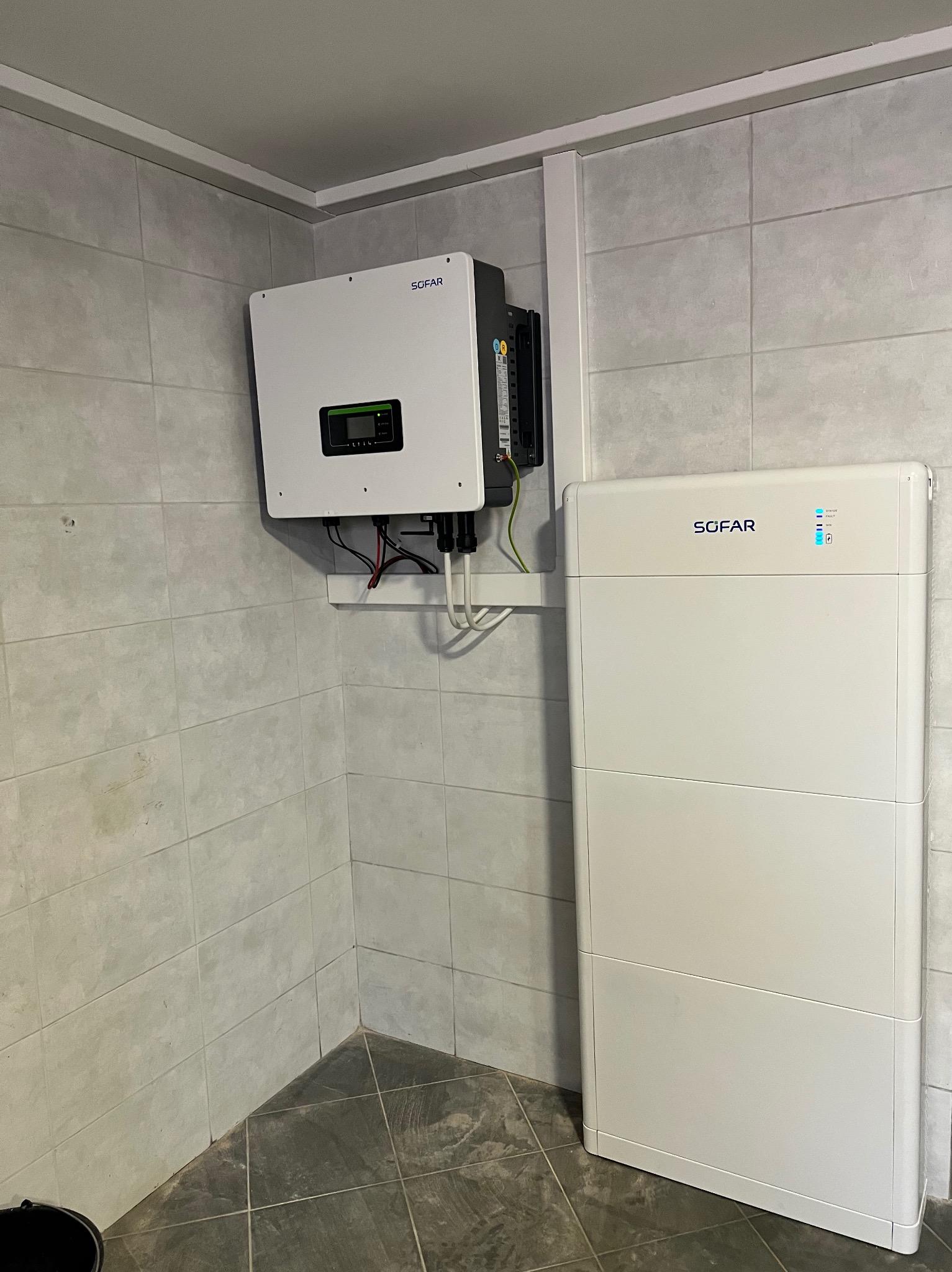 Zamontowany na ścianie biały falownik Sofar z podłączonymi kablami oraz biały akumulator energii Sofar stojący obok w pomieszczeniu z wykafelkowanymi ścianami.