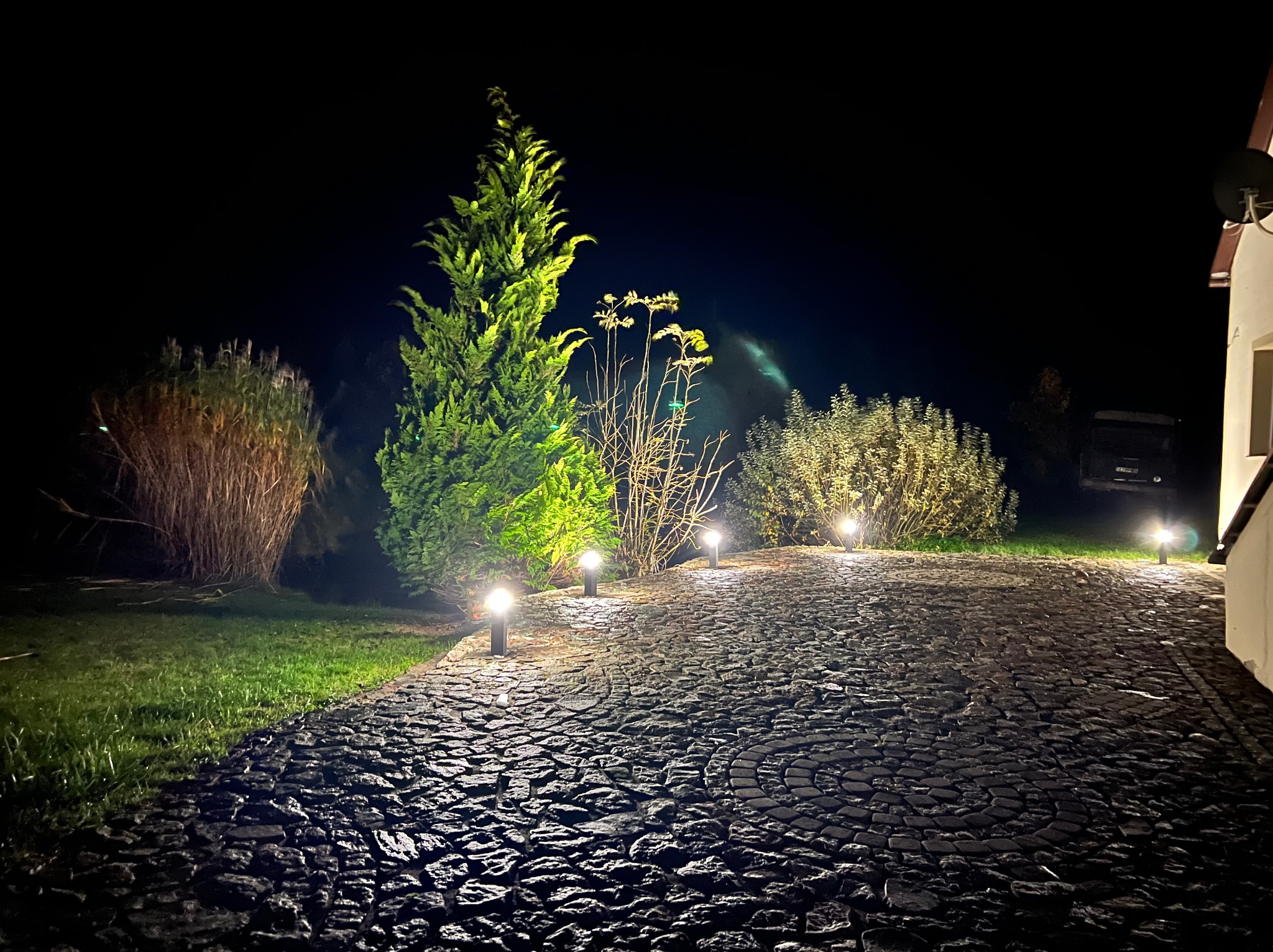 Nocne oświetlenie posesji z lampami najazdowymi wzdłuż kamiennej drogi i podświetlonymi krzewami, tworzące nastrojowy klimat.