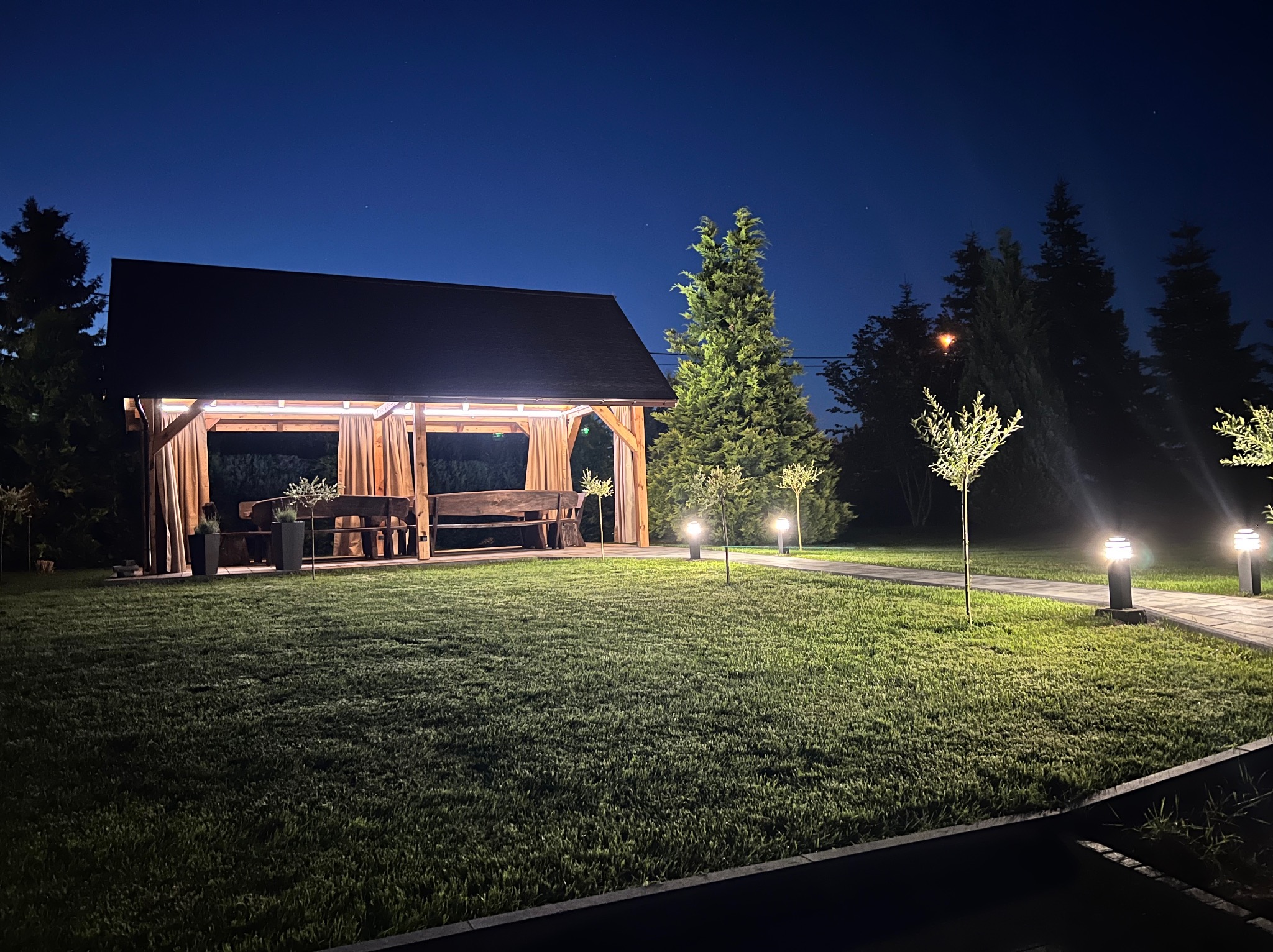 Oświetlona altana z drewnianym stołem i zasłonami w ogrodzie nocą. Ścieżka oświetlona lampami prowadzi przez trawnik.