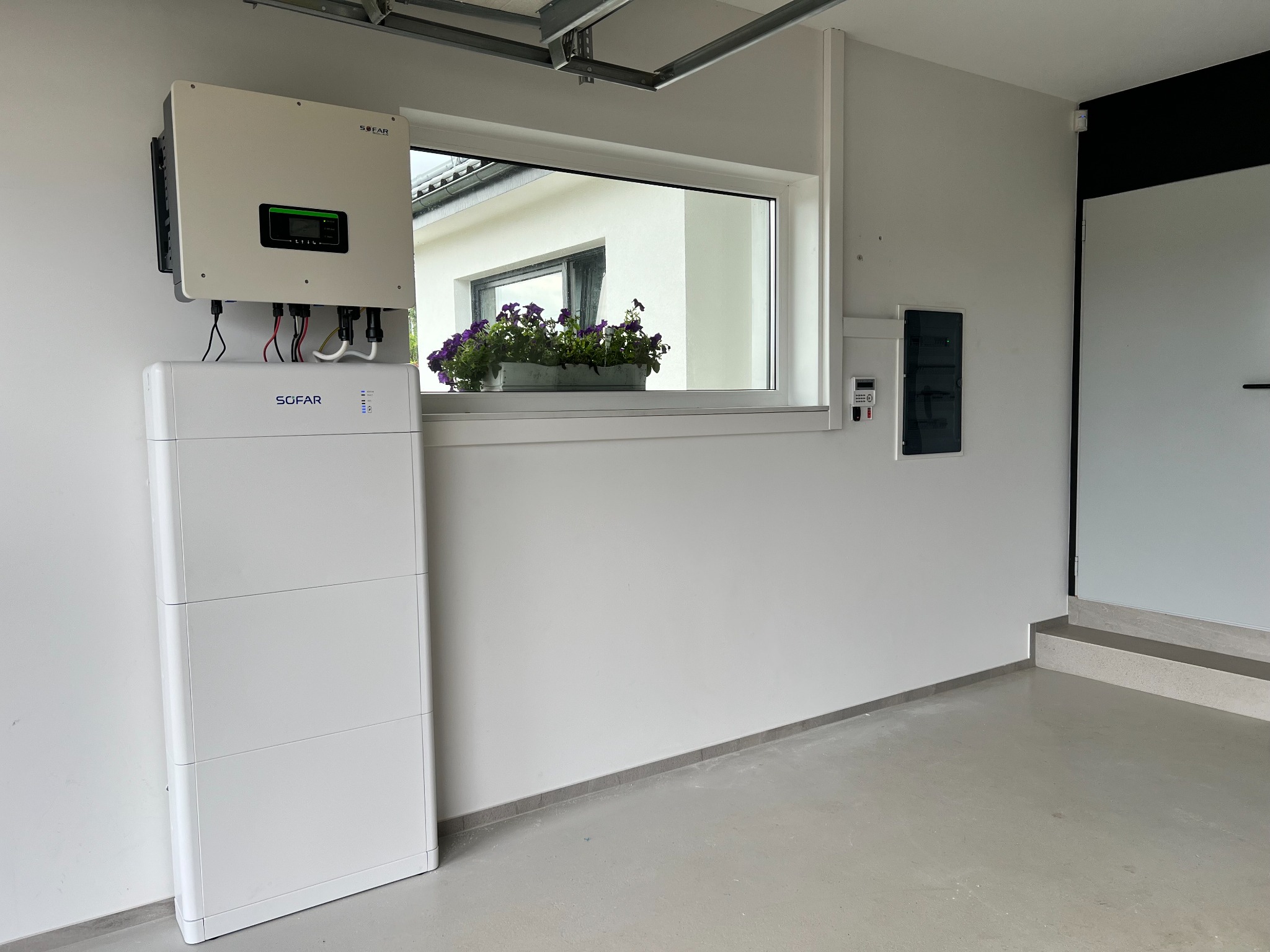 Biały magazyn energii SOFAR z inwerterem na ścianie w garażu, okno z kwiatami, szara podłoga i listwa przypodłogowa. Estetyczny montaż w jasnym wnętrzu.