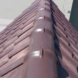 Brązowy dach z dachówki ceramicznej, widok z dołu na kalenicę i fragment elewacji budynku w tle. Widoczne mocowania dachowe.