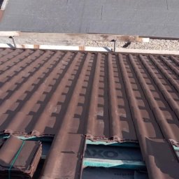 Sz.Til Szymon Thiel - Montaż brązowej dachówki ceramicznej na dachu budynku. Widoczne pakiety dachówek do ułożenia i fragment konstrukcji dachu.