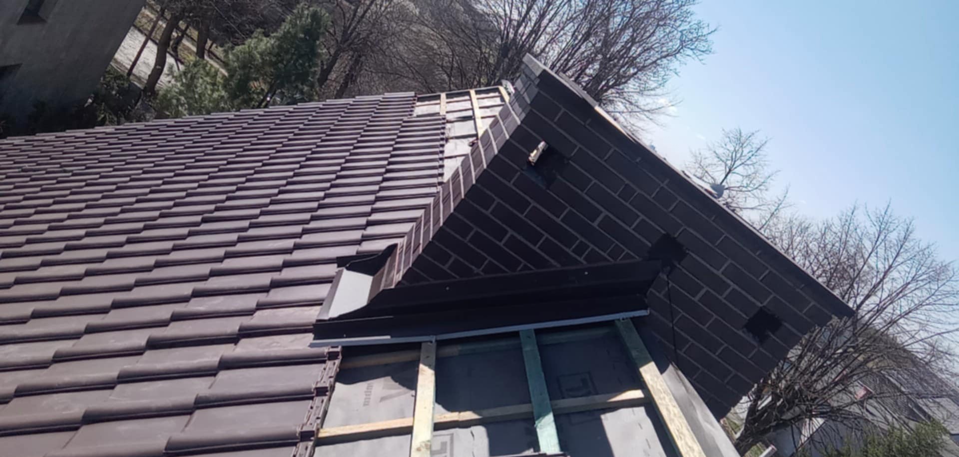 Montaż brązowej dachówki ceramicznej na skośnym dachu, z widoczną konstrukcją wsporczą i elementami obróbki blacharskiej. W tle drzewa i błękitne niebo.