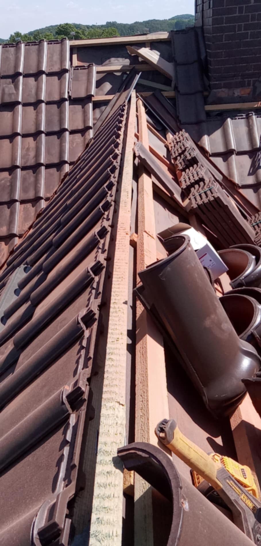 Montaż brązowej dachówki karpiówki na drewnianej konstrukcji dachu, widoczne narzędzia i elementy obróbki blacharskiej. Kąt ujęcia od dołu.