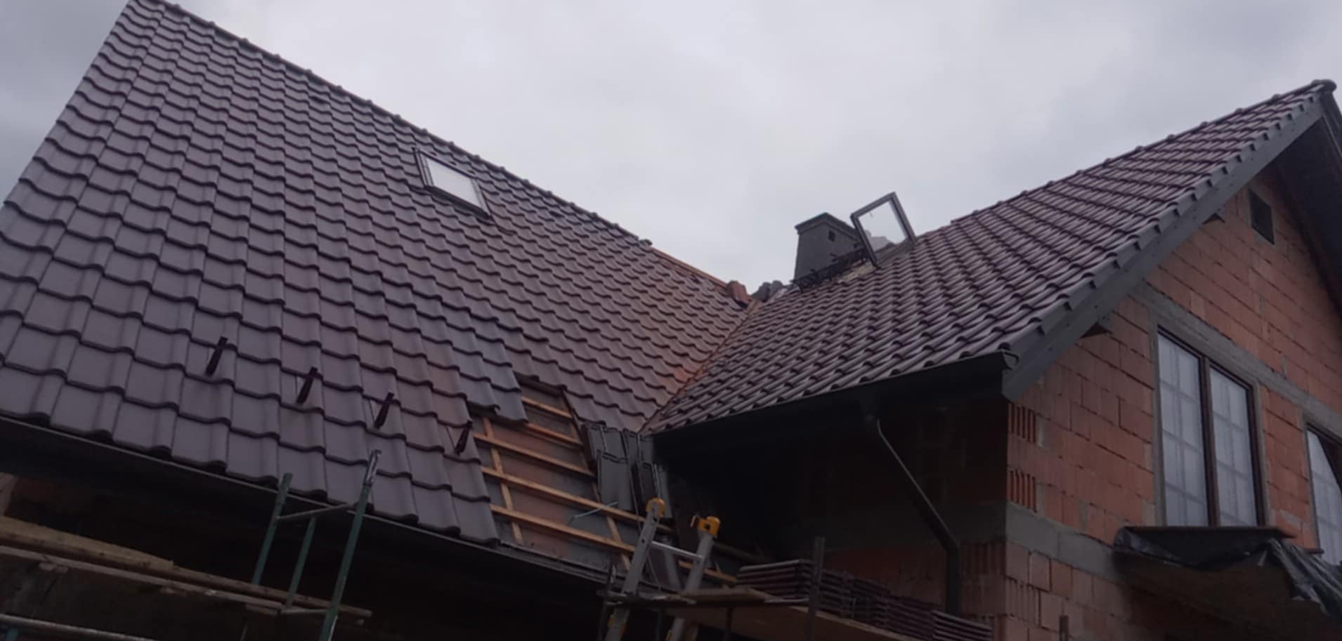 Montaż brązowej dachówki na skośnym dachu domu jednorodzinnego z oknem dachowym i kominem; widoczne rusztowanie i fragment niezakończonej części dachu.