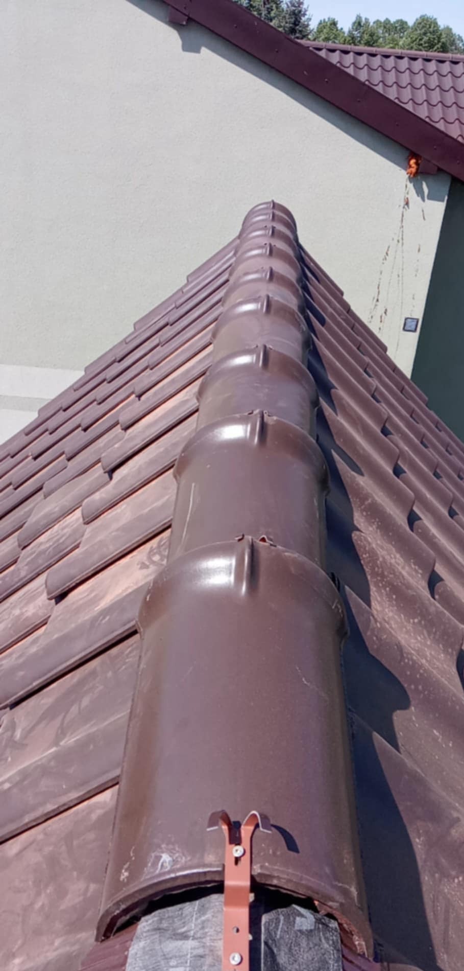 Brązowy dach z dachówki ceramicznej, widok z dołu na kalenicę i fragment elewacji budynku w tle. Widoczne mocowania dachowe.