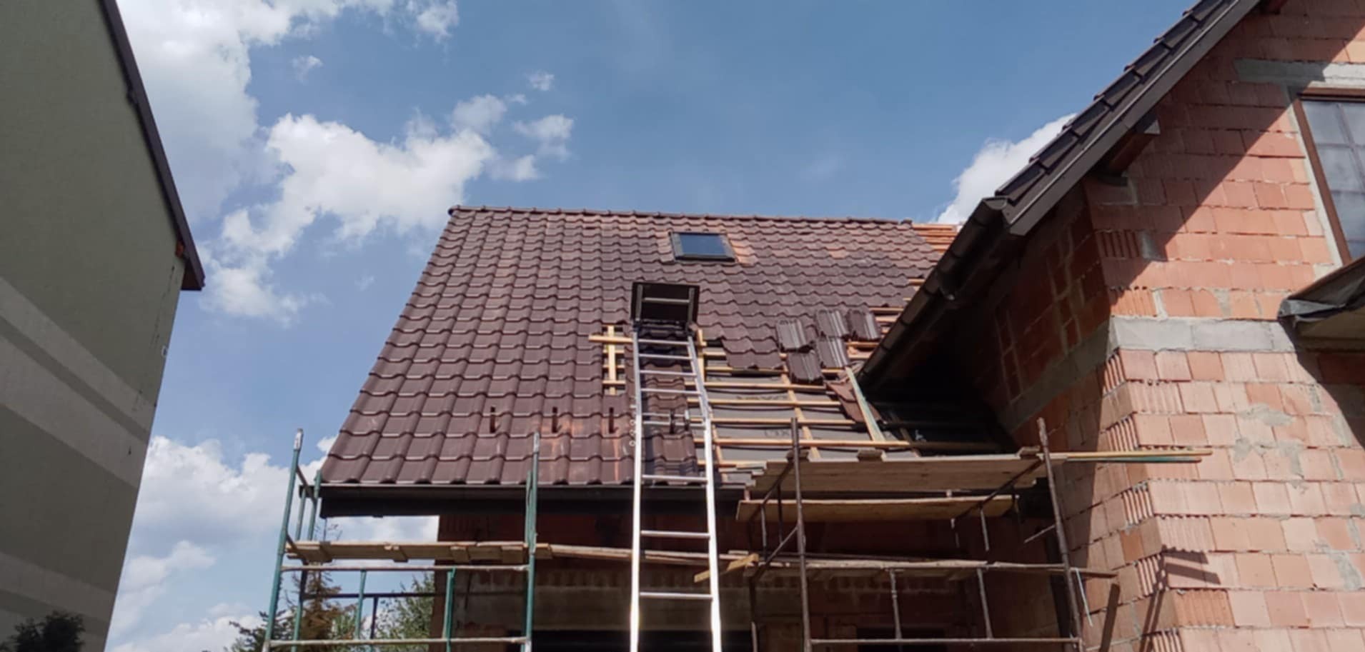 Ceglany dom w budowie z częściowo wykończonym, brązowym dachem z dachówką. Na dachu drabina i rusztowanie, widoczne niebo z chmurami.
