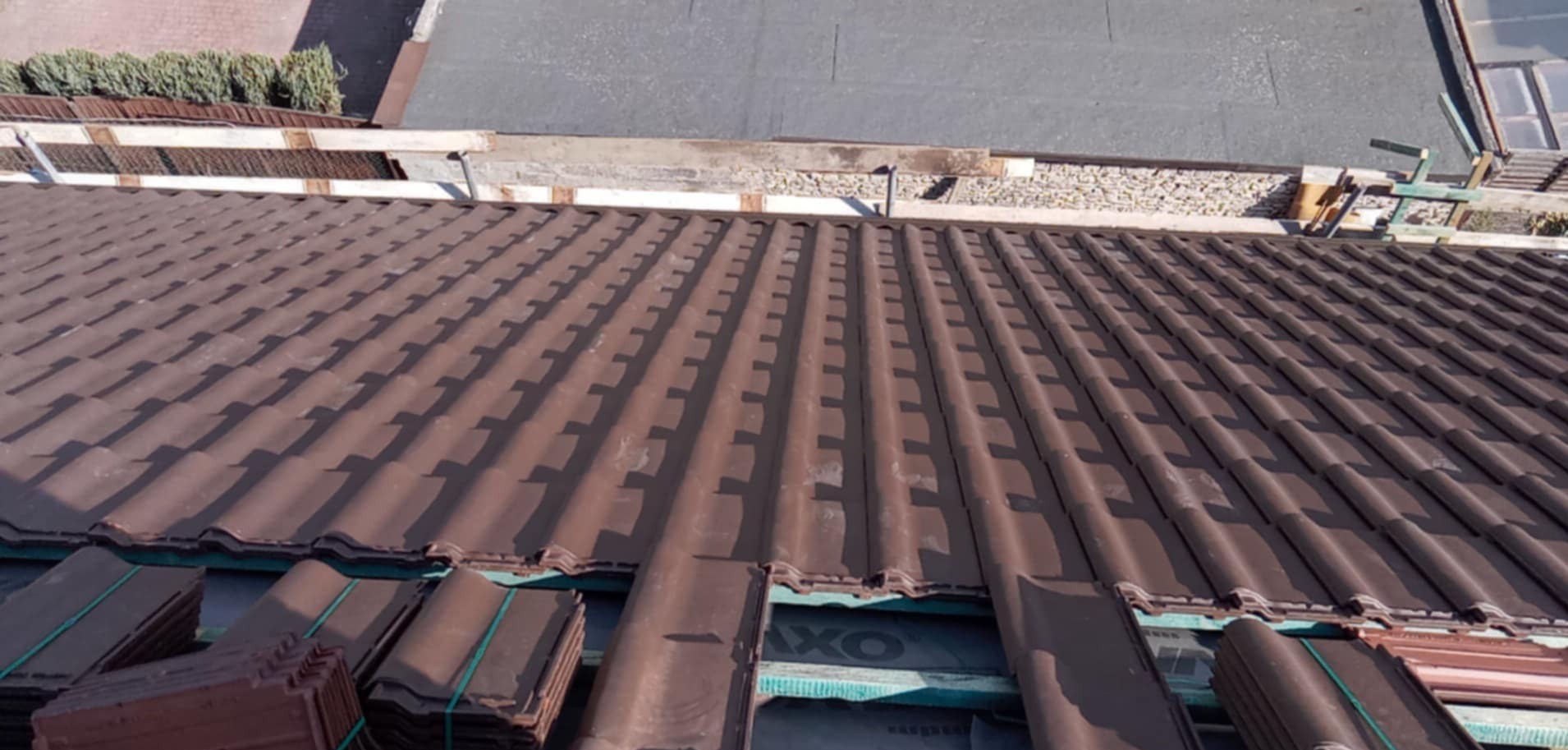 Montaż brązowej dachówki ceramicznej na dachu budynku. Widoczne pakiety dachówek do ułożenia i fragment konstrukcji dachu.