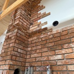 Marcin Zalewski - Ceglana ściana w trakcie wykończenia z widoczną drabiną i drewnianym stropem. Częściowo położona cegła dekoracyjna na białej ścianie.