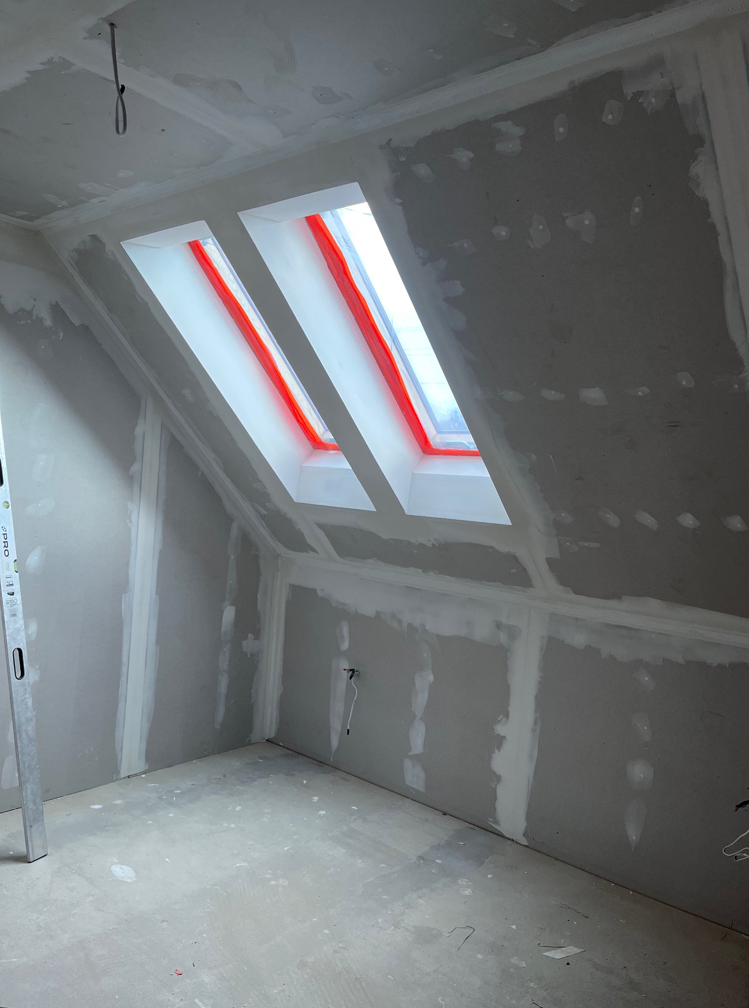 Pokój na poddaszu w trakcie szpachlowania, widoczne dwa okna dachowe z czerwonym zabezpieczeniem, ściany przygotowane do malowania, surowy beton na podłodze.