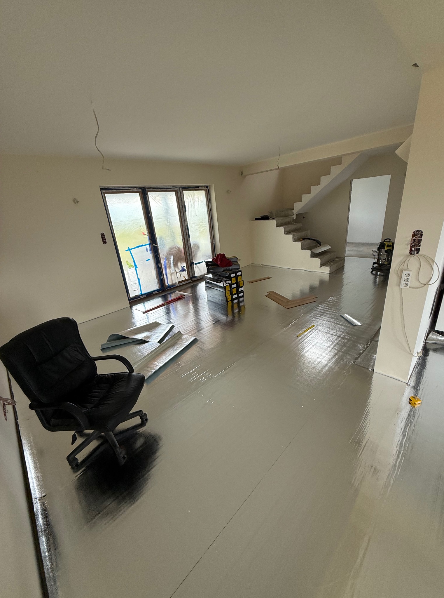 Przygotowanie do układania paneli: podkład izolacyjny na podłodze, widoczne panele, narzędzia i schody w tle. Okno zabezpieczone folią.