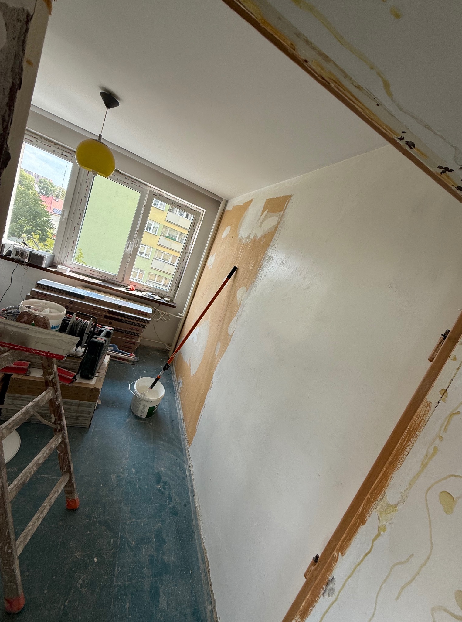 Pokój w trakcie malowania: wałek oparty o ścianę, wiadro z farbą, drabina, okno z widokiem na budynek. Fragment ściany pomalowany na biało, reszta przygotowana do malowania.
