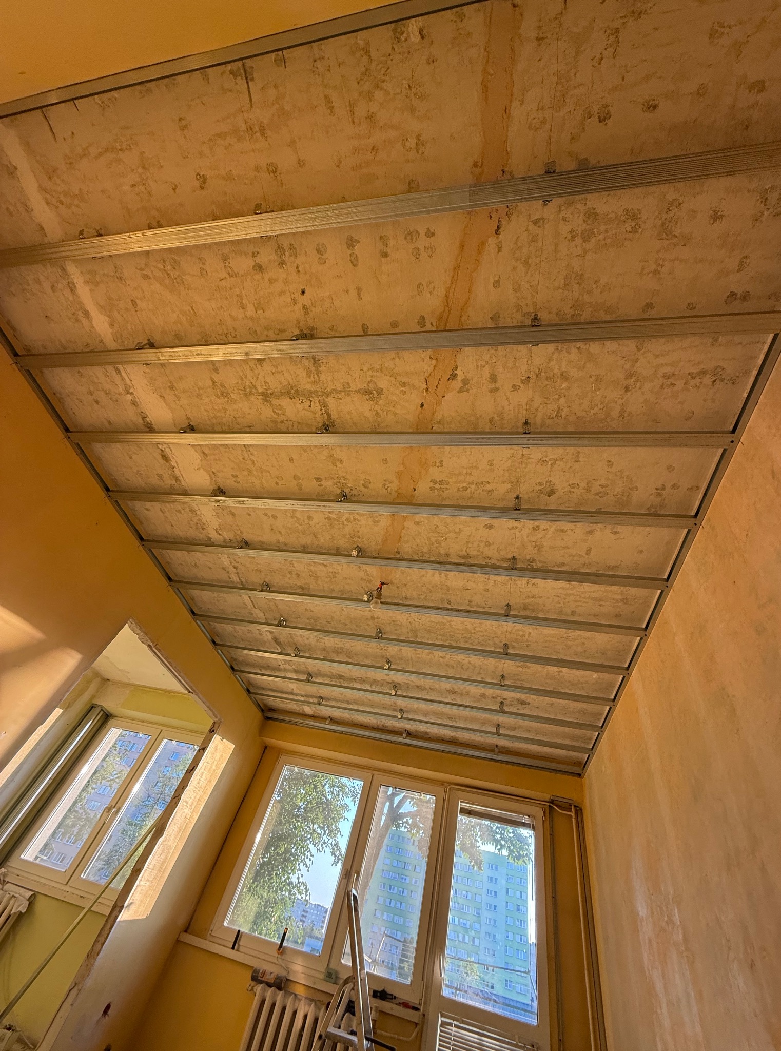 Montaż stelażu sufitu podwieszanego w pokoju o żółtych ścianach, widoczne okna z widokiem na zewnątrz. Na podłodze drabina. Prace wykończeniowe w trakcie.