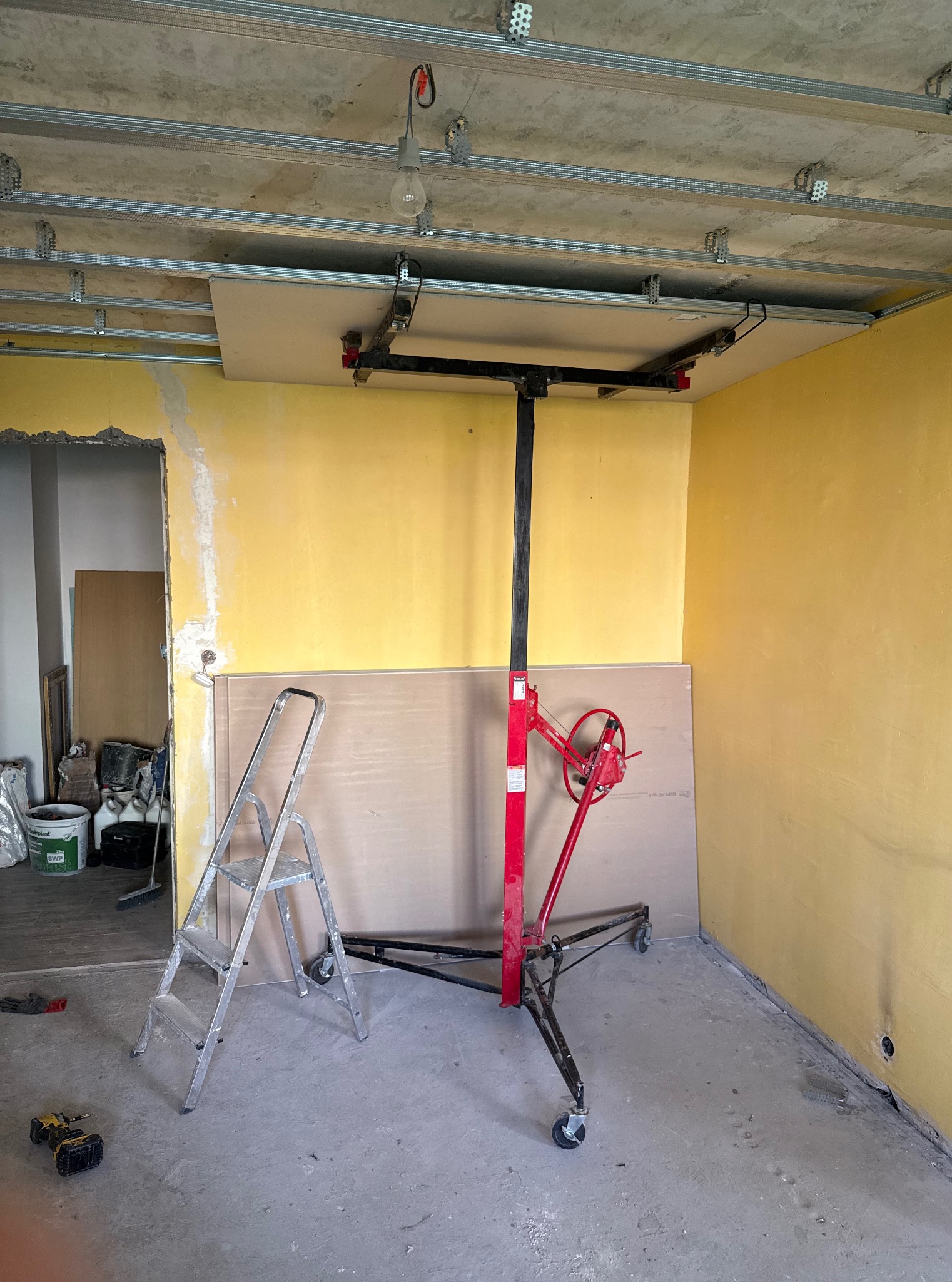 Żółty pokój w trakcie montażu sufitu podwieszanego z płyt gipsowo-kartonowych. Widoczny podnośnik do płyt i drabina. Częściowo wykończone ściany.