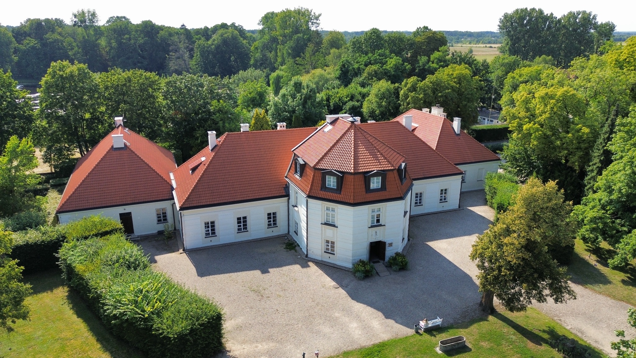 Biały dwór z czerwoną dachówką, otoczony zielenią, widziany z lotu ptaka w słoneczny dzień. Charakterystyczna wieża z oknami i geometryczne dachy.