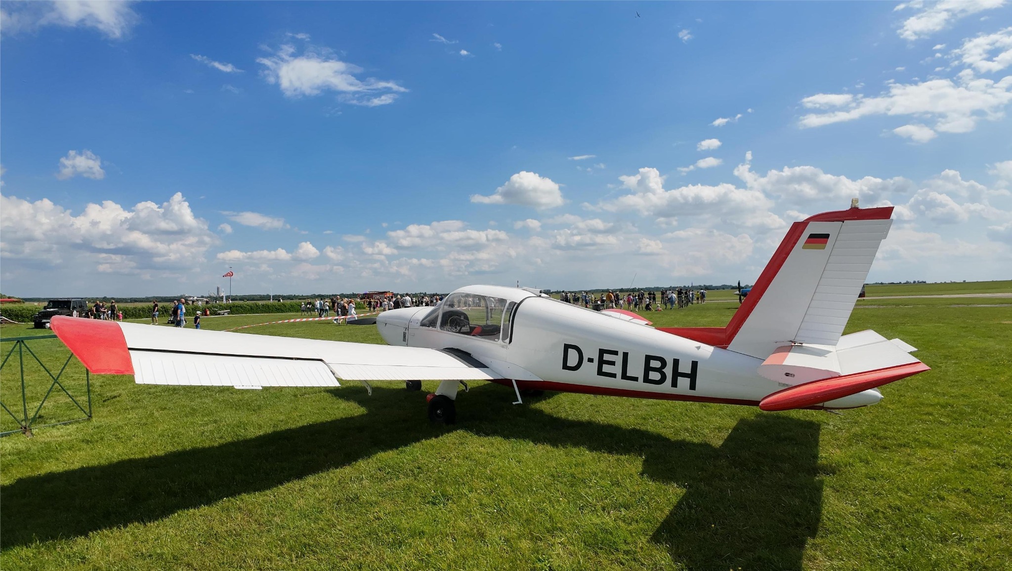 Biało-czerwony samolot na trawiastym lotnisku, w tle grupa ludzi i błękitne niebo z chmurami. Widoczny znak rejestracyjny D-ELBH i flaga Niemiec na stateczniku pionowym.