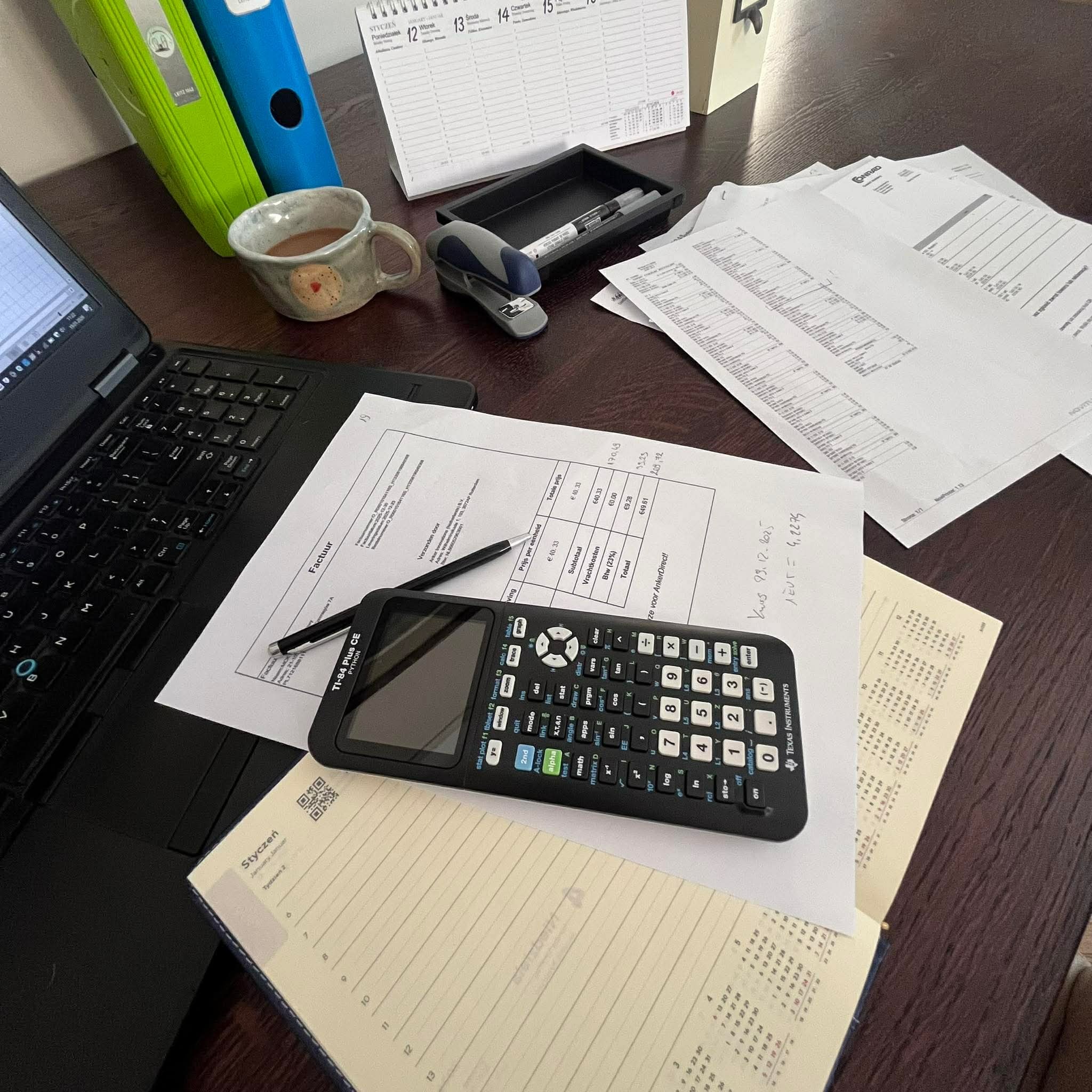 Biurko z kalkulatorem, fakturą, kalendarzem i laptopem. Elementy pracy biurowej w kadrze, sugerujące usługi finansowe lub księgowe. Widok z góry, oświetlenie dzienne.