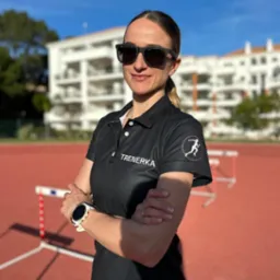 Uśmiechnięta trenerka w okularach na bieżni, ubrana w koszulkę polo z napisem 'TRENERKA' i logo biegacza, z budynkiem w tle.