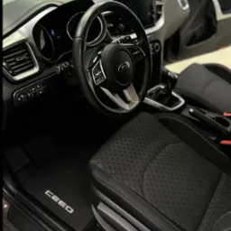 Wnętrze samochodu Kia Ceed z czarną tapicerką i gumowymi dywanikami z napisem CEED. Widoczna kierownica, deska rozdzielcza i otwarte drzwi.