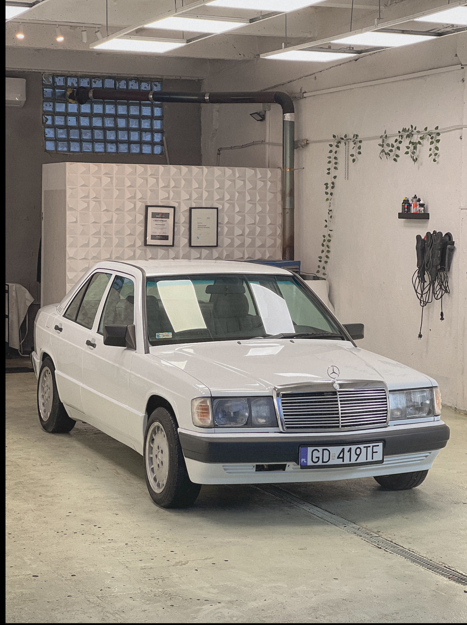 Biały Mercedes-Benz 190 w warsztacie z certyfikatami na ścianie. Widoczny grill, reflektory, tablica rejestracyjna i elementy dekoracyjne wnętrza warsztatu.