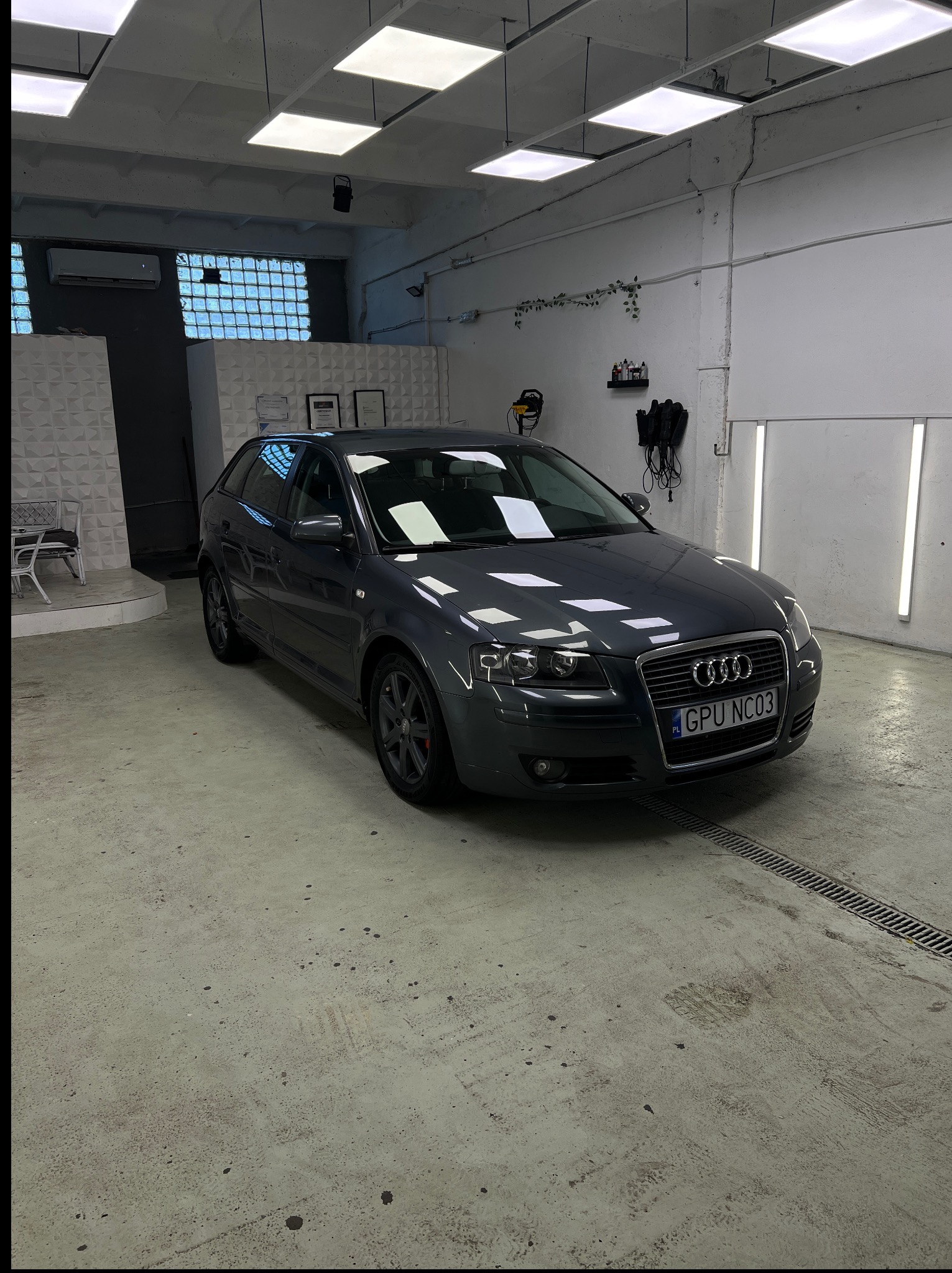Szary Audi A3 po myciu w jasnym garażu z kratką ściekową w podłodze, widoczne oświetlenie sufitowe i narzędzia na ścianie. Tablica rejestracyjna GPU NC03.