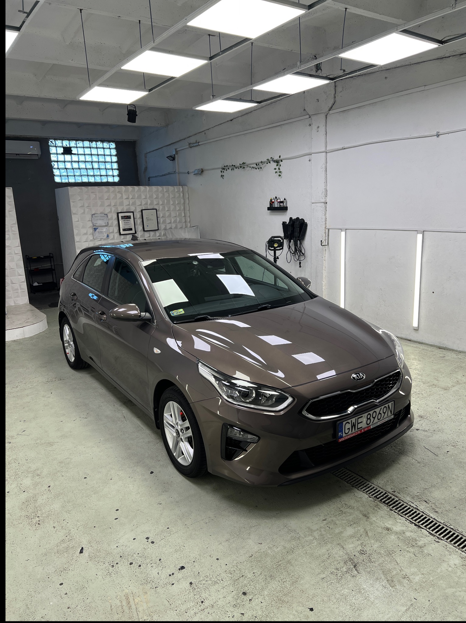 Brązowa Kia Ceed po myciu w jasnym pomieszczeniu myjni, z widocznymi certyfikatami na ścianie i kratką odpływową w posadzce. Oświetlenie sufitowe podkreśla czystość.
