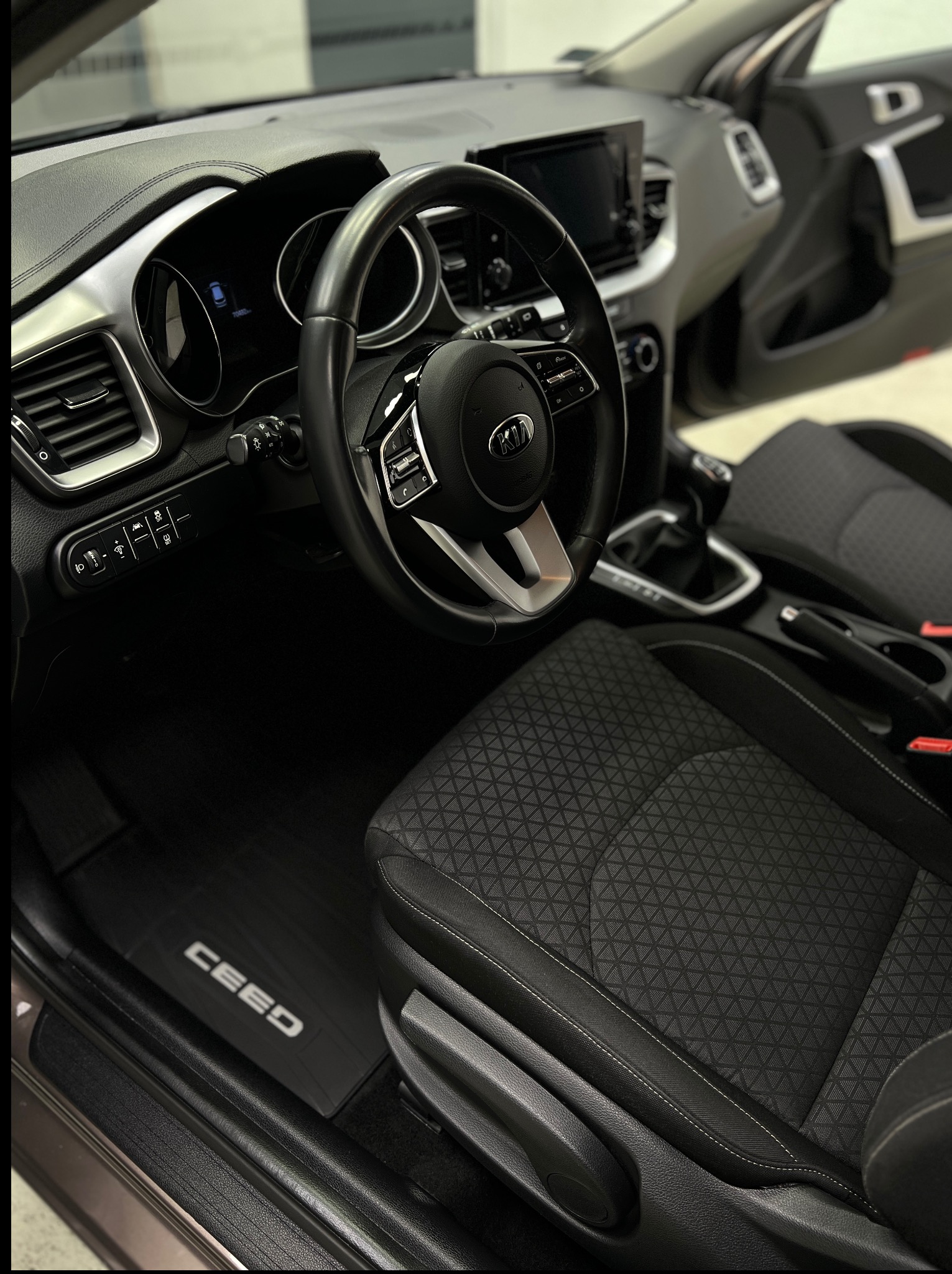 Wnętrze samochodu Kia Ceed z czarną tapicerką i gumowymi dywanikami z napisem CEED. Widoczna kierownica, deska rozdzielcza i otwarte drzwi.