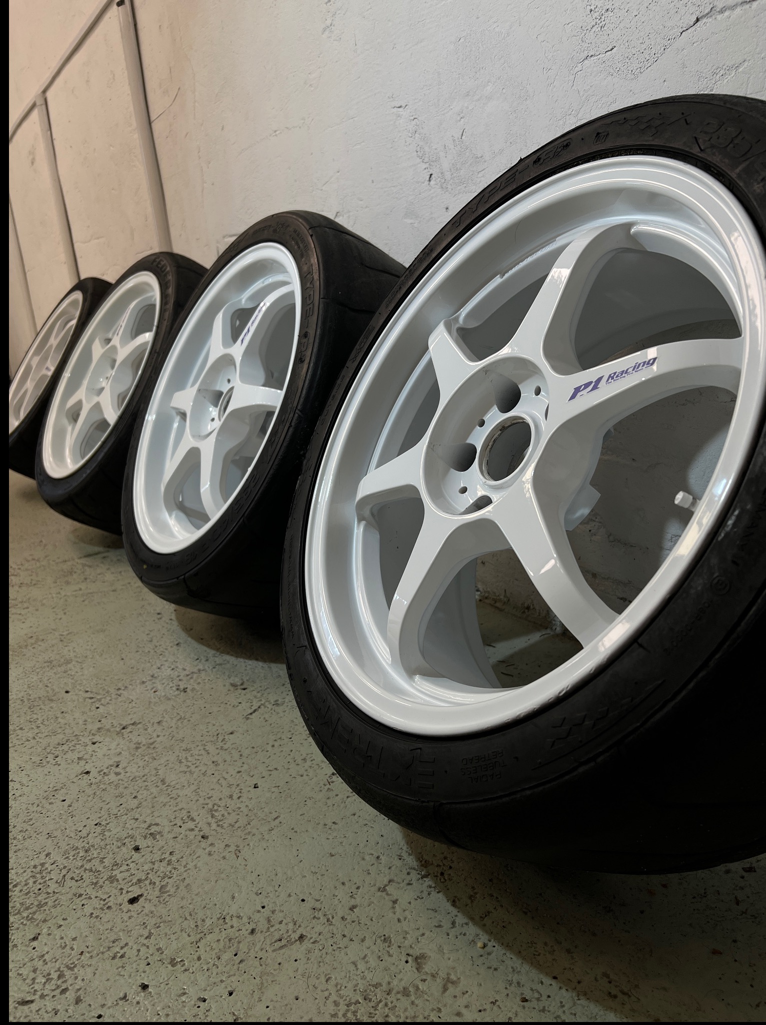 Rząd czterech białych felg z oponami P1 Racing ustawionych pod kątem na szarym betonowym podłożu w garażu. Widoczne ślady użytkowania opon.