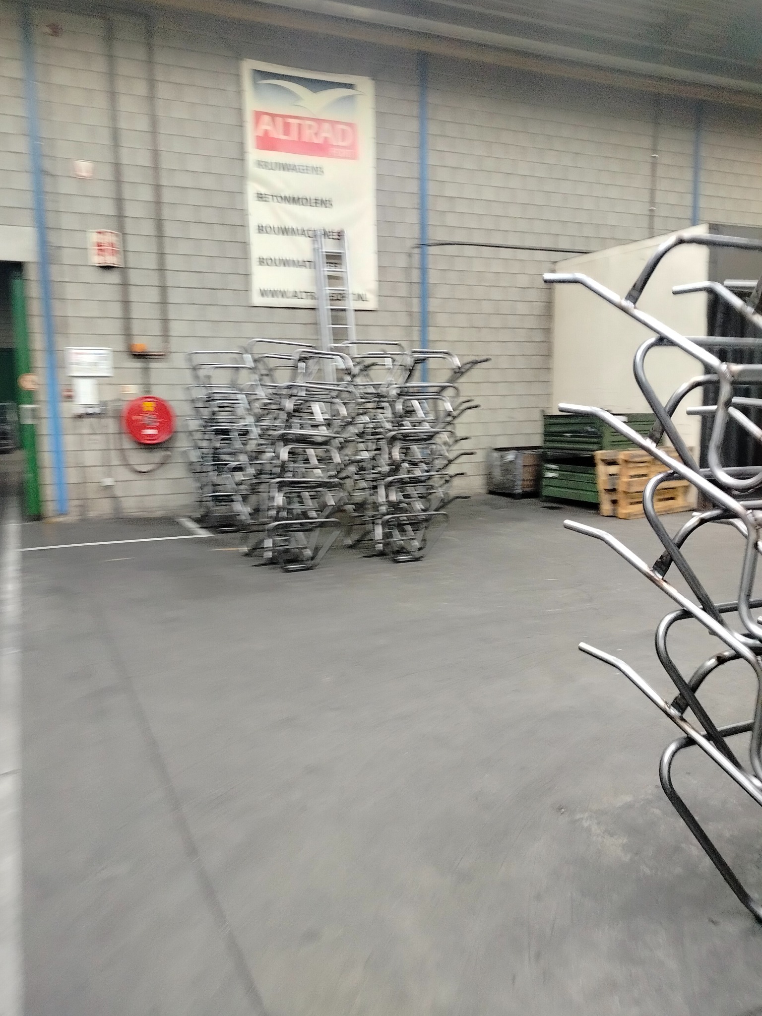 Stos metalowych ram w hali produkcyjnej, z widocznym logo firmy Altrad na ścianie w tle. Ramy ustawione w rzędach, tworzą regularny wzór. Widoczny sprzęt gaśniczy.