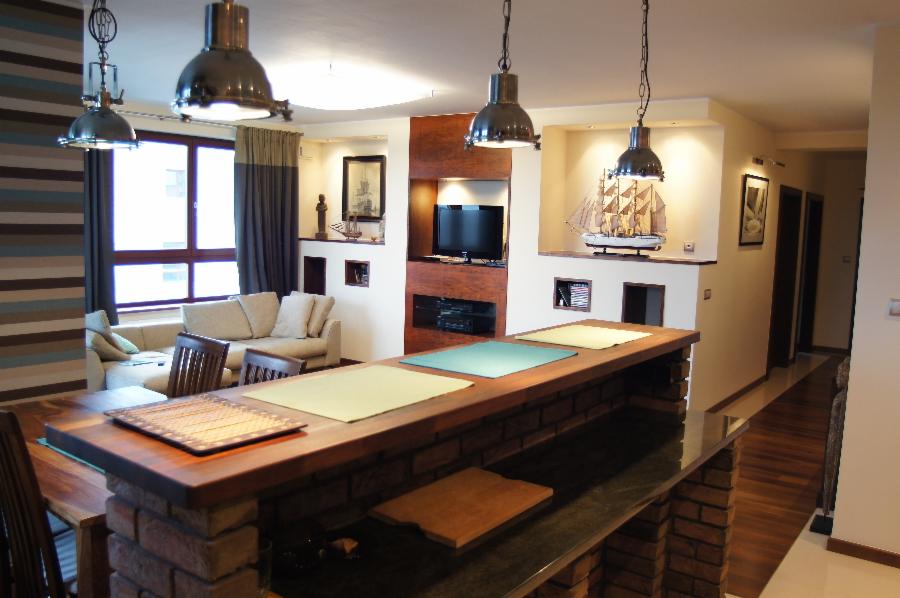 Salon z ceglanym barkiem i marynistycznymi dekoracjami. Widoczny fragment sofy, stół z krzesłami i oświetlenie w stylu industrialnym. Aranżacja wnętrza w jasnych barwach.