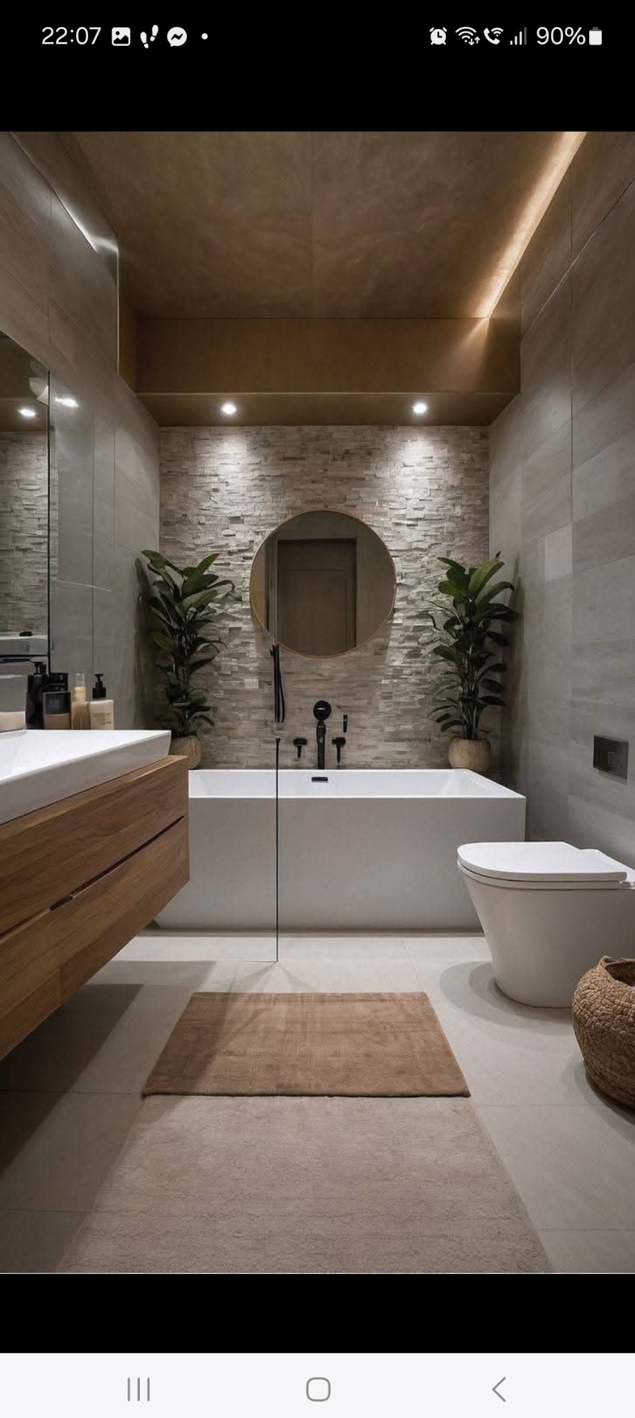 Nowoczesna łazienka z kamienną ścianą, okrągłym lustrem nad wanną, drewnianą szafką pod umywalką i roślinami w donicach. Stonowana kolorystyka.