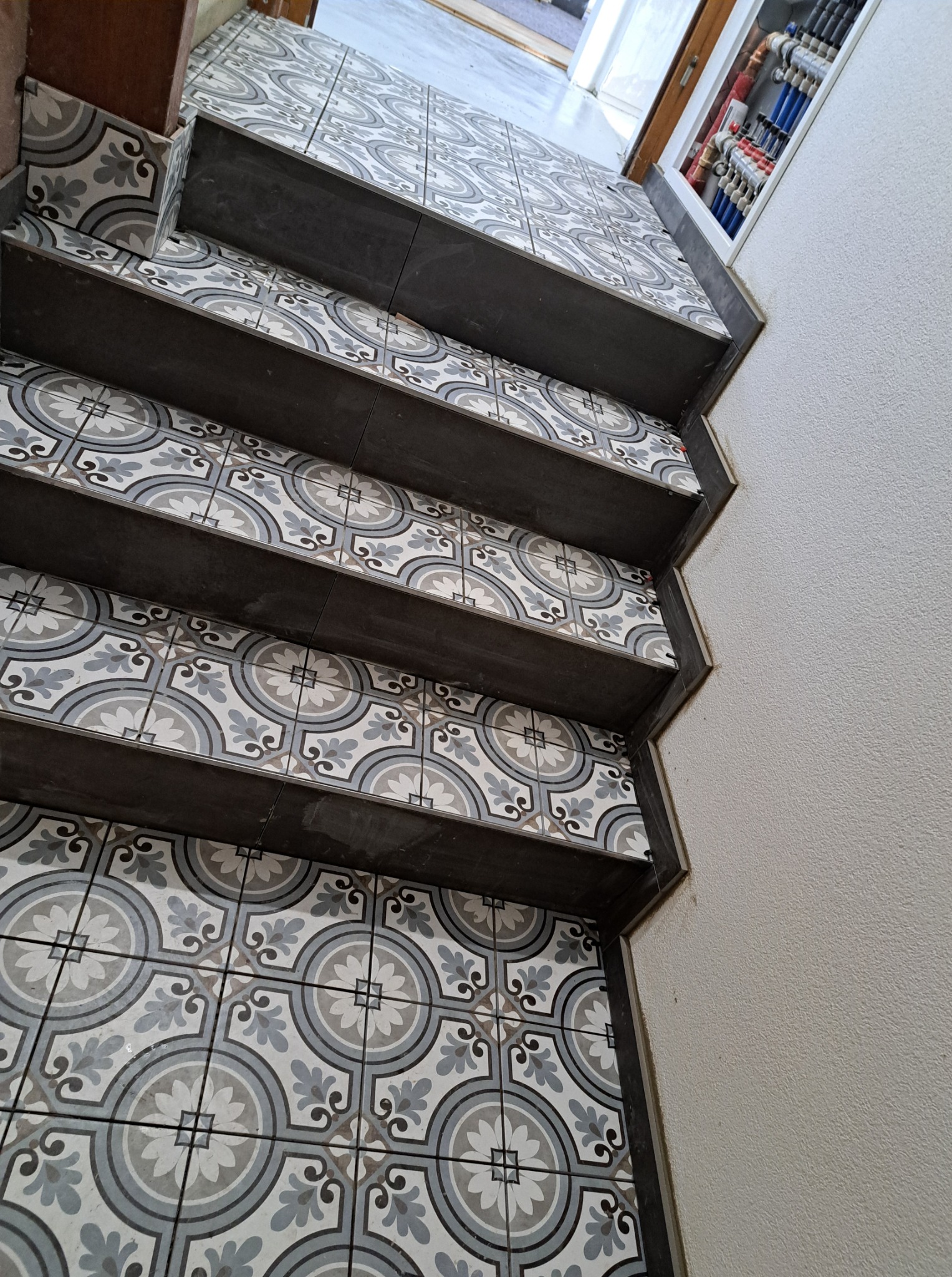 Wykończone schody z dekoracyjnymi, szaro-białymi płytkami. Widoczne rury instalacji w tle. Nietypowy kąt ujęcia podkreśla wzór płytek i staranność wykonania.