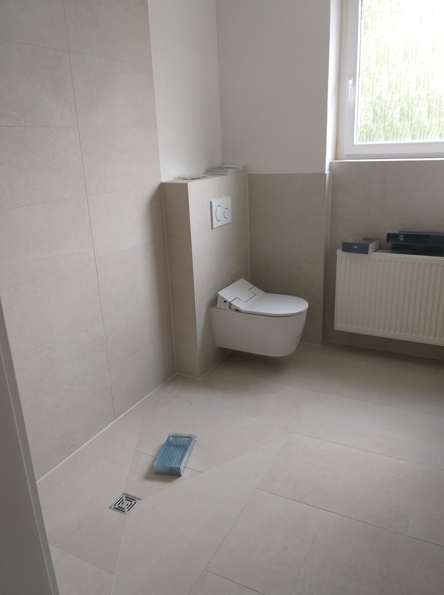 Jasna łazienka po remoncie: beżowe płytki na ścianach i podłodze, wisząca toaleta, grzejnik pod oknem i odpływ liniowy z kratką. Minimalistyczny design, czystość i nowoczesność.