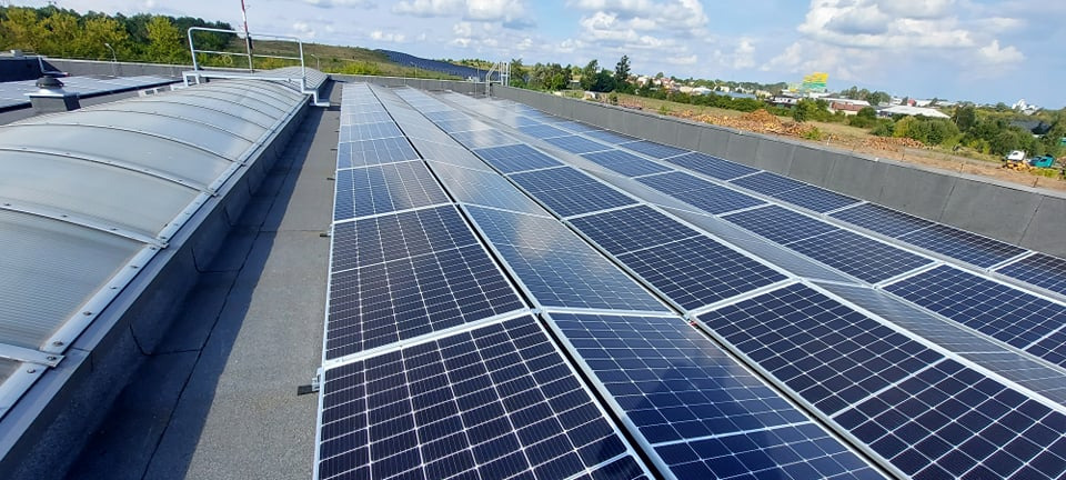 Rząd paneli fotowoltaicznych na płaskim dachu budynku z widocznym świetlikiem i barierkami. W tle zabudowa miejska i błękitne niebo z chmurami.