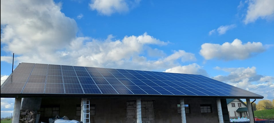 Panele słoneczne na dachu budynku gospodarczego, błękitne niebo z chmurami w tle. Widoczna drabina oparta o ścianę budynku.