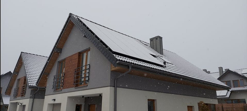 Dom jednorodzinny z panelami słonecznymi na dachu, pokryty świeżym śniegiem. Widok z dołu, pochmurne niebo. Zima.